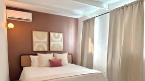 Ein Schlafzimmer mit einem Bett, einer Klimaanlage, zwei Wandbildern und einem Fenster mit Vorhängen.