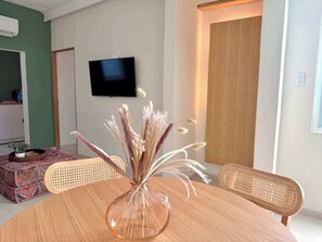 A modern living room with a round table, a clear glass vase with dried pampas grass, a television on the wall, and a wooden door. - Sea Sun & Pool - Charming 1BR Apt (San Juan)