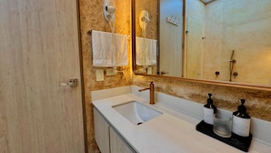 Un baño con pared beige texturizada, un lavabo blanco, un grifo dorado y un espejo sobre el lavabo. Hay dos botellas de crema de manos en la encimera.