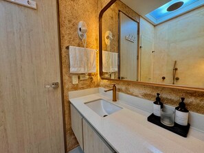 Un baño con pared beige texturizada, un lavabo blanco, un grifo dorado y un espejo sobre el lavabo. Hay dos botellas de crema de manos en la encimera.
