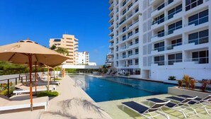 Zona de piscina con sillones de descanso y sombrillas, rodeada de edificios modernos.