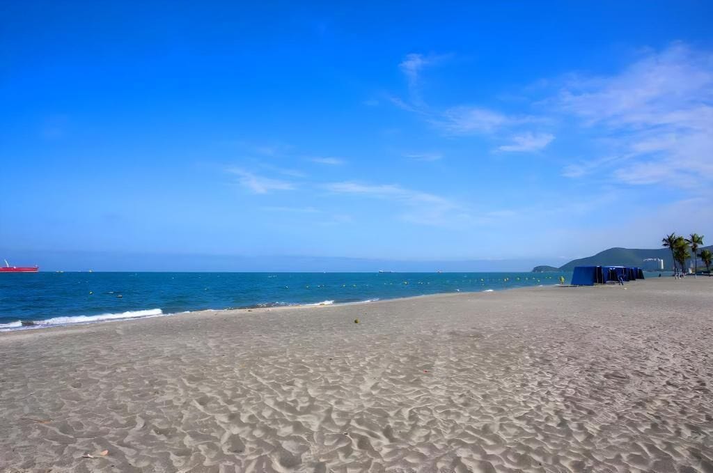 Une plage avec un ciel dégagé, une mer calme et quelques tentes visibles au loin.