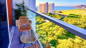 A balcony with wicker chairs, a glass door, and a view of a cityscape and greenery. - Suite Vista A Reserva y Mar Piso Alto En Bello Horizonte (Santa Marta)