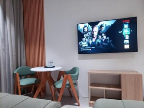 Une chambre d’hôtel moderne avec un téléviseur à écran plat fixé au mur, deux fauteuils verts autour d’une petite table ronde et une table d’appoint en bois avec une étagère.