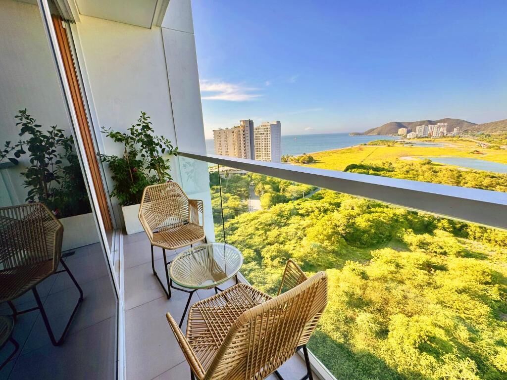 Un balcón con sillas de mimbre y una mesa, con vistas a un paisaje costero con edificios y vegetación.