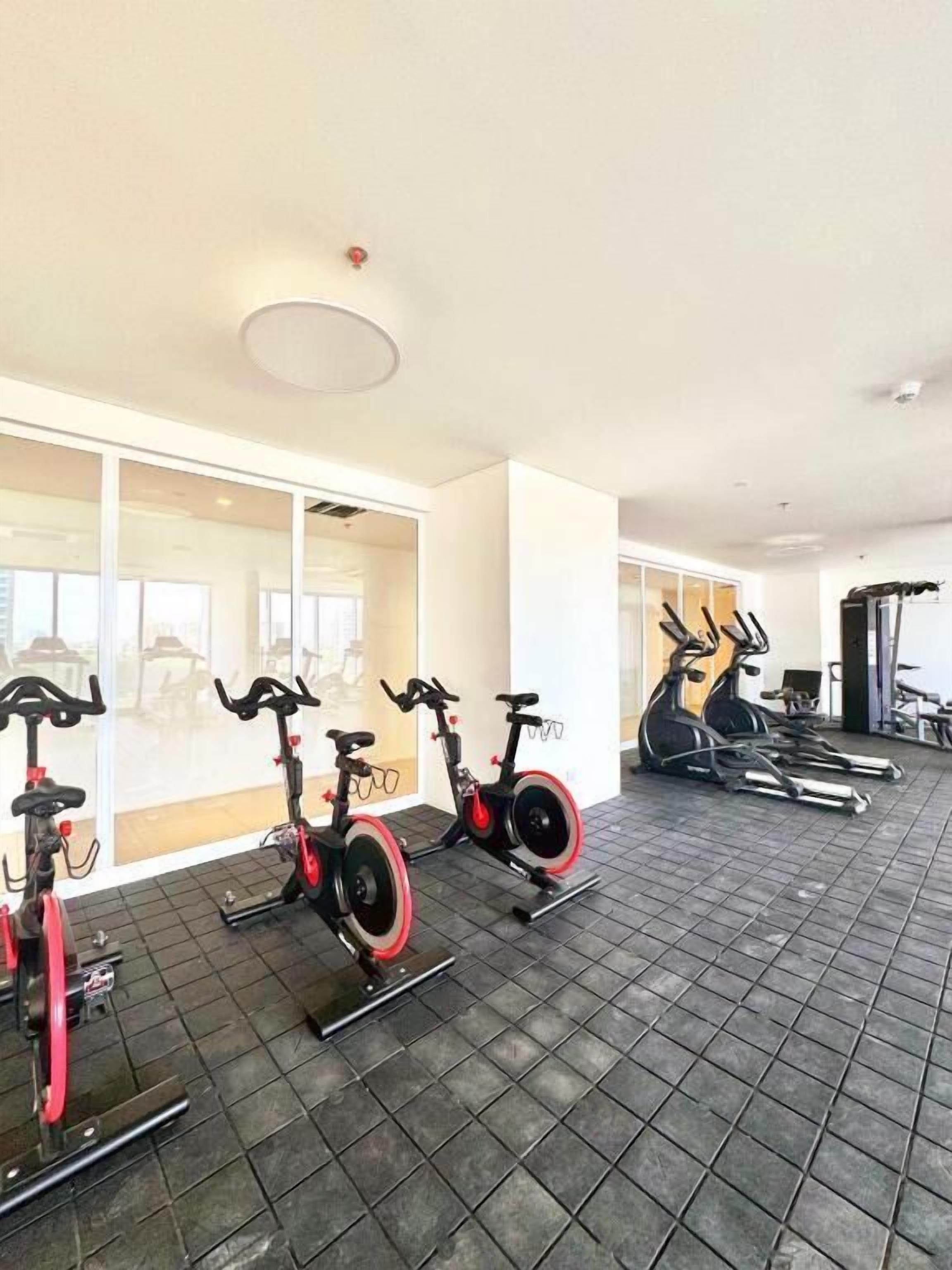 Un gimnasio con bicicletas estáticas y cintas de correr, ventanas amplias y una lámpara en el techo.