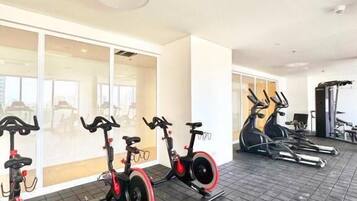 Un gimnasio con bicicletas estáticas y cintas de correr, ventanas amplias y una lámpara en el techo.