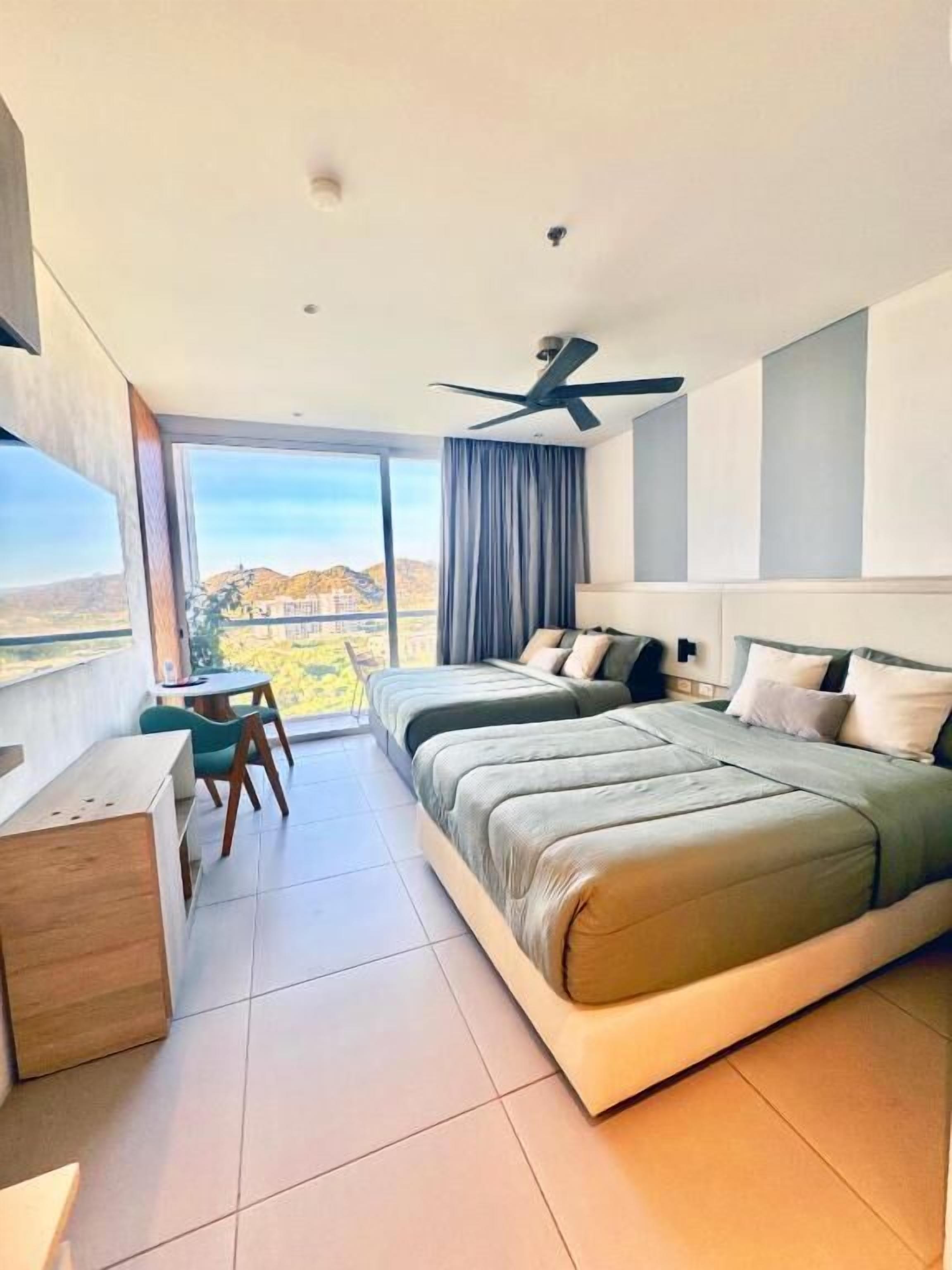 Una habitación de hotel moderna con una cama grande, un escritorio, una silla, un ventilador de techo y vistas a un paisaje montañoso.