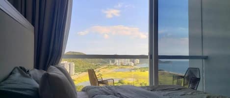 A bedroom with a large window, a bed, a chair, and a view of a cityscape and hills.