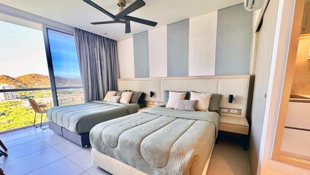 A modern hotel room with two beds, a large window with a view, a ceiling fan, and a balcony. - Suite Vista A Reserva y Mar Piso Alto En Bello Horizonte (Santa Marta)