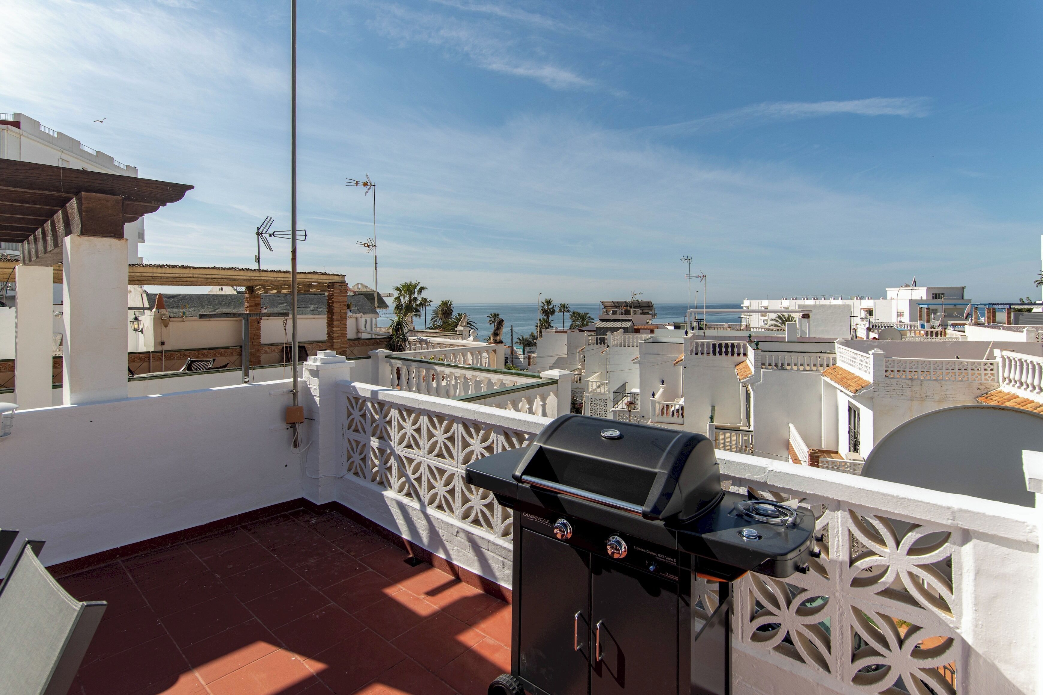 A rooftop terrace with a barbecue grill, white railings, and a view of the sea and buildings.