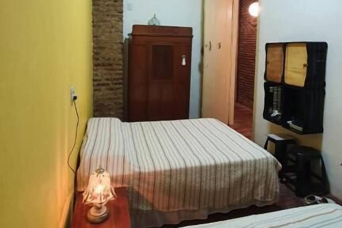 A bedroom with two beds, a wooden ceiling, a stone wall, a wardrobe, and a small table with a lamp.