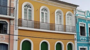 A yellow building with white-framed windows and green doors, a balcony with metal railings, and a neighboring blue building. - Hostel Casa de Avany (Cachoeira)