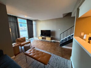 A modern living room with a flat-screen TV, leather sofa, ottoman, and a staircase. - Great 2x1 Loft Apartment in Downtown Austin (Austin)
