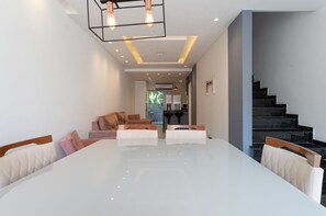 Uma sala de jantar moderna com uma mesa branca grande, cadeiras estofadas, uma escada e uma cozinha ao fundo.