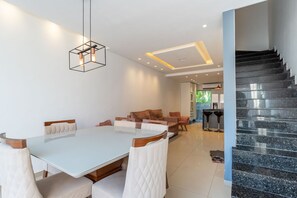Uma área de refeições moderna, com mesa de vidro e cadeiras estofadas brancas, um sofá de couro marrom e uma escada preta.