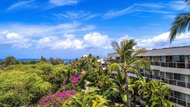 A hotel with a lush garden, palm trees, and a view of the ocean. - Aston Maui Banyan H302 | Pool & Hot Tub (Kihei)