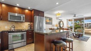 A modern kitchen with stainless steel appliances, a large island with bar stools, and a dining area with a view. - Aston Maui Banyan H302 | Pool & Hot Tub (Kihei)
