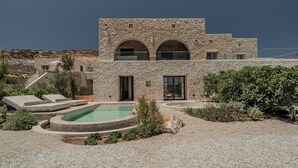 A modern stone building with a swimming pool, outdoor seating, and a view of the surrounding landscape. - Eunoia Suites (Paros)