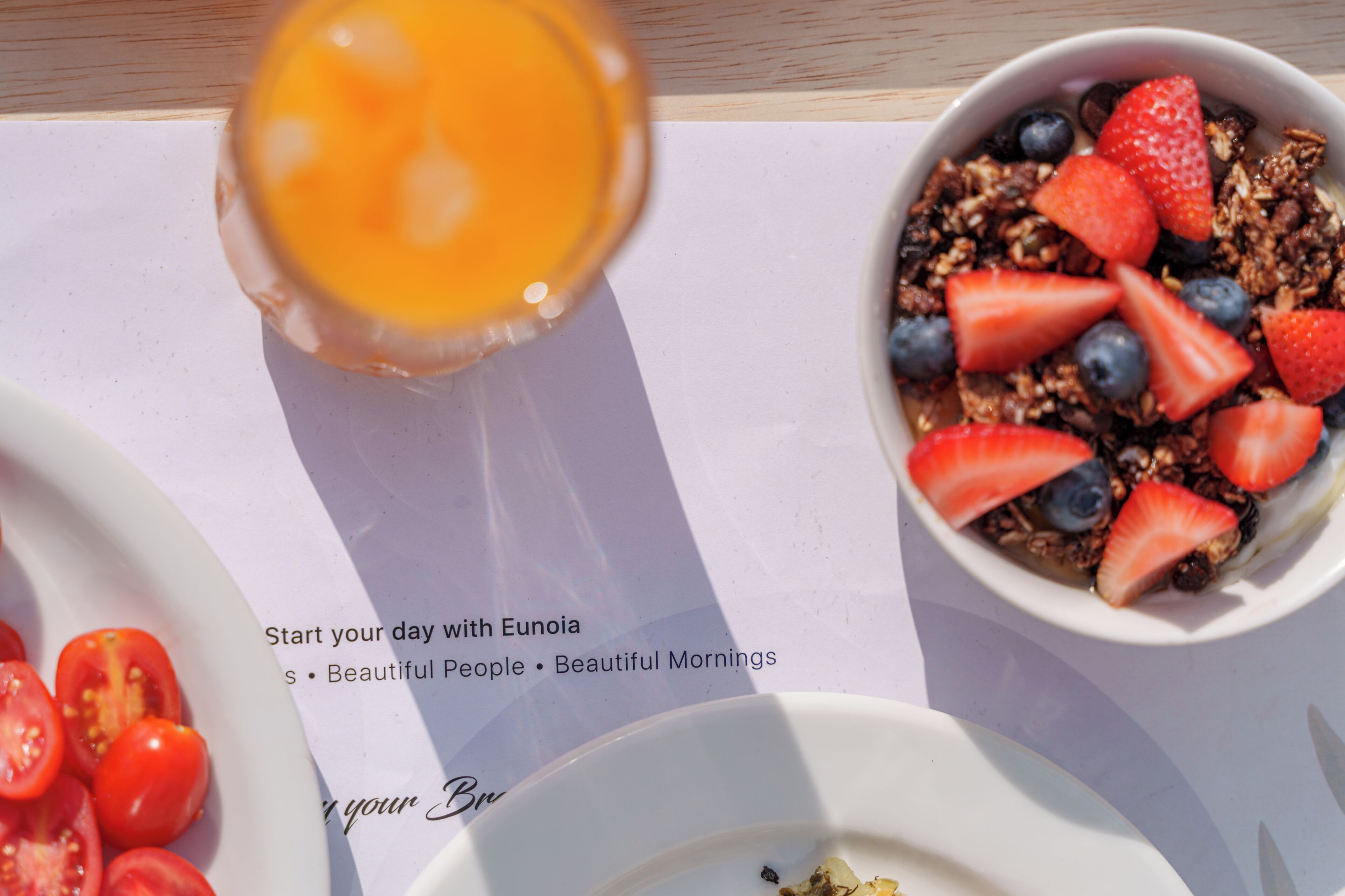 A breakfast setting with a bowl of granola, strawberries, and blueberries, a glass of orange juice, and a plate of sliced tomatoes.