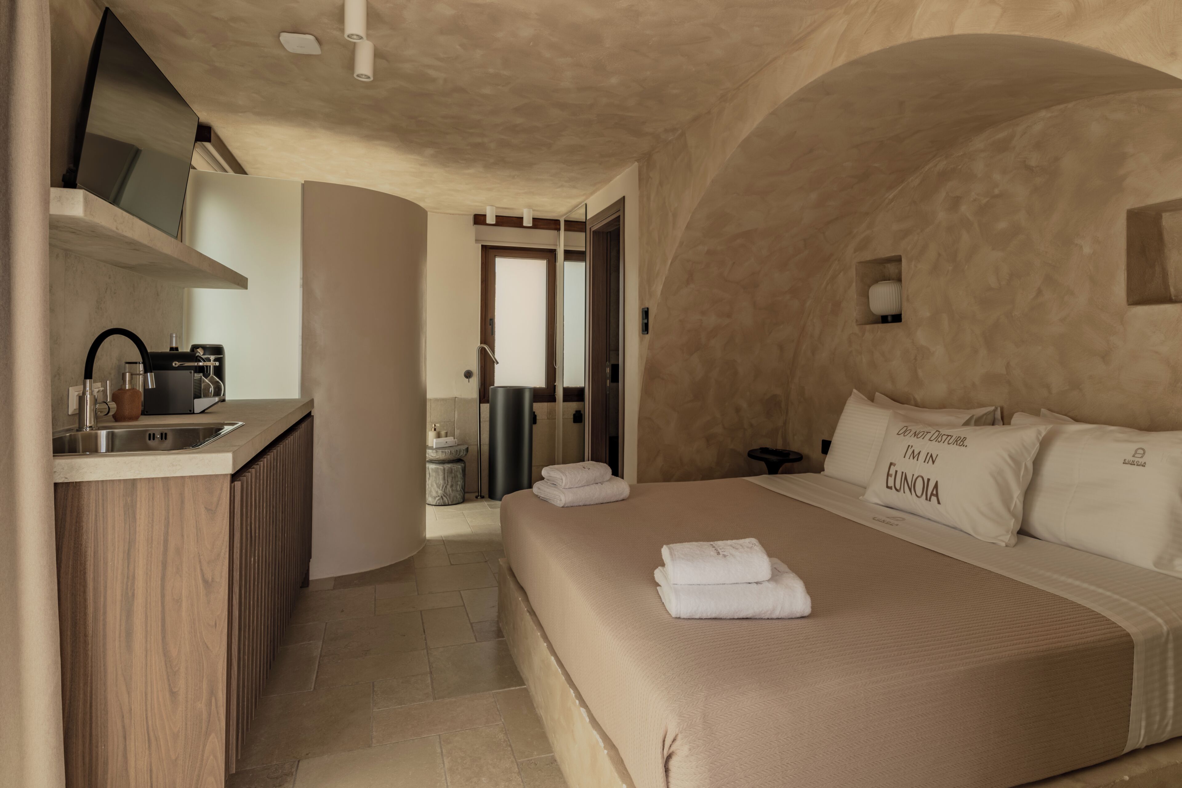 A modern hotel room with a kitchenette, a large bed with white linens, and a unique arched ceiling.