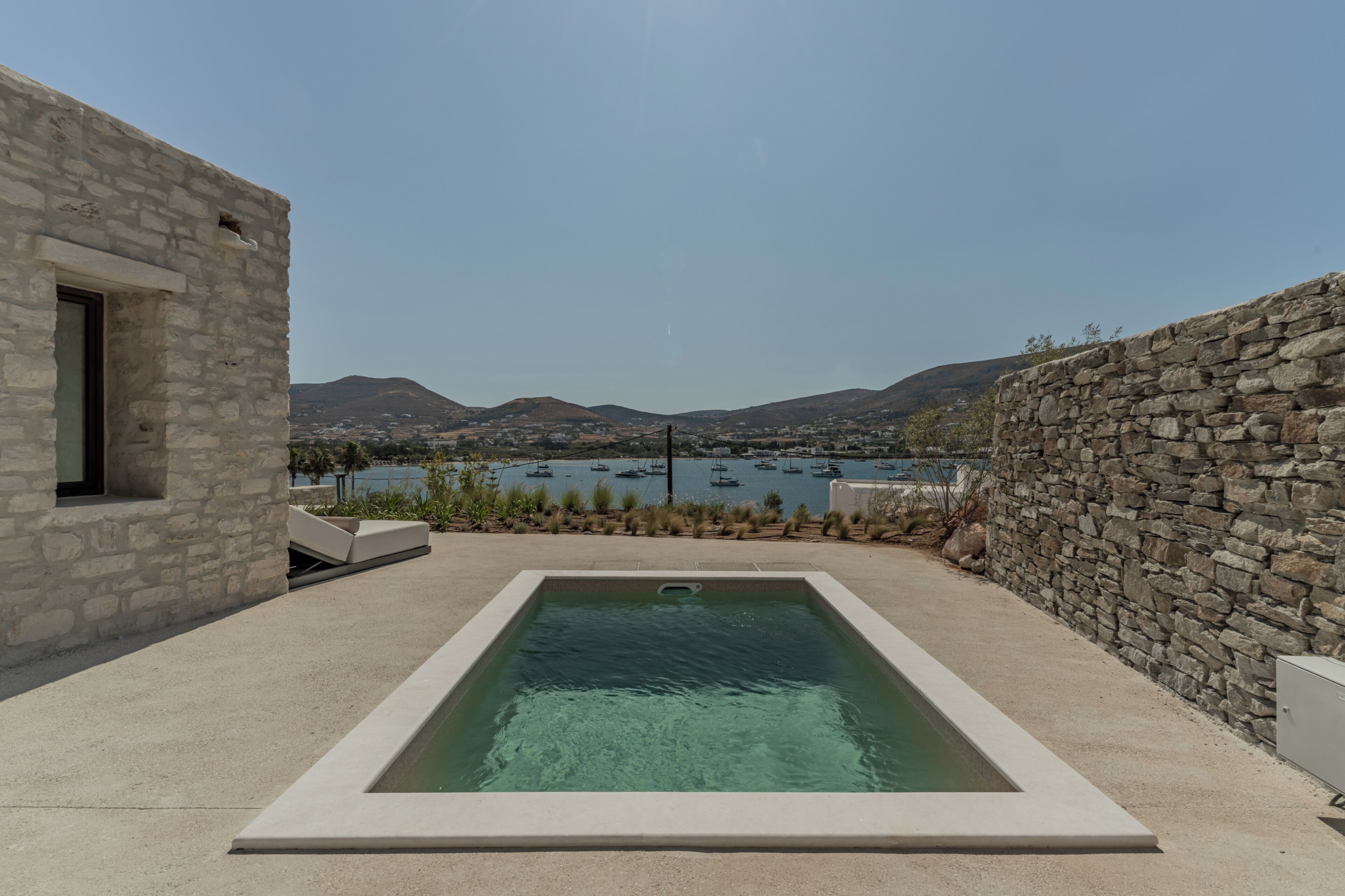A swimming pool with a view of boats on a lake, surrounded by stone walls and a clear sky.