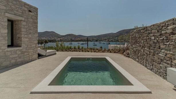 A swimming pool with a view of boats on a lake, surrounded by stone walls and a clear sky. - Eunoia Suites (Paros)