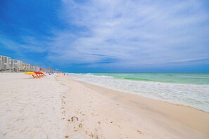 Ein Strand mit einer Reihe von Liegestühlen und Sonnenschirmen, einem sandigen Ufer und einem klaren, blauen Himmel.