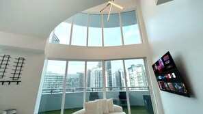 Ein modernes Zimmer mit gewölbter Decke, großen Fenstern, Blick auf die Stadt, einem weißen Sofa und einem an der Wand montierten Fernseher.