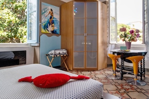A bedroom with a bed, a red pillow, a wardrobe, a table with yellow chairs, and a window with a view of greenery.