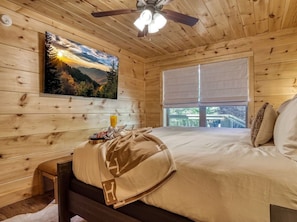 Ein Schlafzimmer einer Holzhütte mit einem Bett, einem Nachttisch, einem Fenster und einem Deckenventilator.