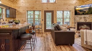 A wooden cabin interior with a kitchen, dining area, and living room, featuring large windows and a stone fireplace. - Twilight Escape | Designer Cabin + Firepit & Views (Sevierville)