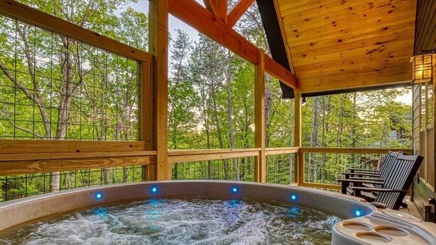 A hot tub with a wooden deck and chairs, surrounded by trees. - Twilight Escape | Designer Cabin + Firepit & Views (Sevierville)
