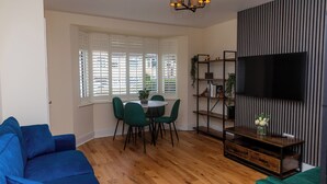 A living room with a blue sofa, a dining table with chairs, a TV on a black wall, and a wooden floor. - Luxury 2 bed house (Bristol)
