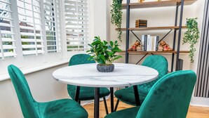 Une table ronde entourée de quatre chaises de couleur turquoise, une plante en pot et une bibliothèque remplie de livres et d’objets décoratifs.