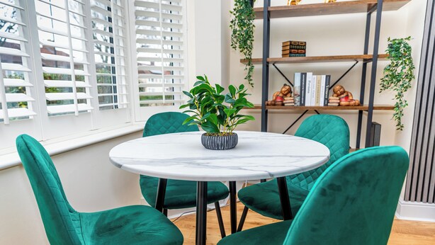 A round table with four teal chairs, a potted plant, and a bookshelf with books and decorative items. - Luxury 2 bed house (Bristol)