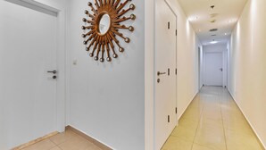 A hallway with white walls, a sunburst mirror, and a tiled floor. - bnbme - Emirates Crown - 2BR & Maid Room (Dubai)