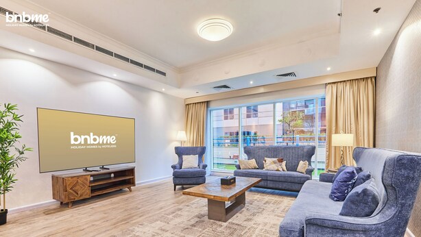 A modern living room with a flat-screen TV, sofas, a coffee table, and a large window with curtains. - bnbme - Emirates Crown - 2BR & Maid Room (Dubai)