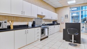 A modern kitchen with white cabinets, a stove, and a microwave. There is a bar stool and a window with a view. - bnbme - Emirates Crown - 2BR & Maid Room (Dubai)