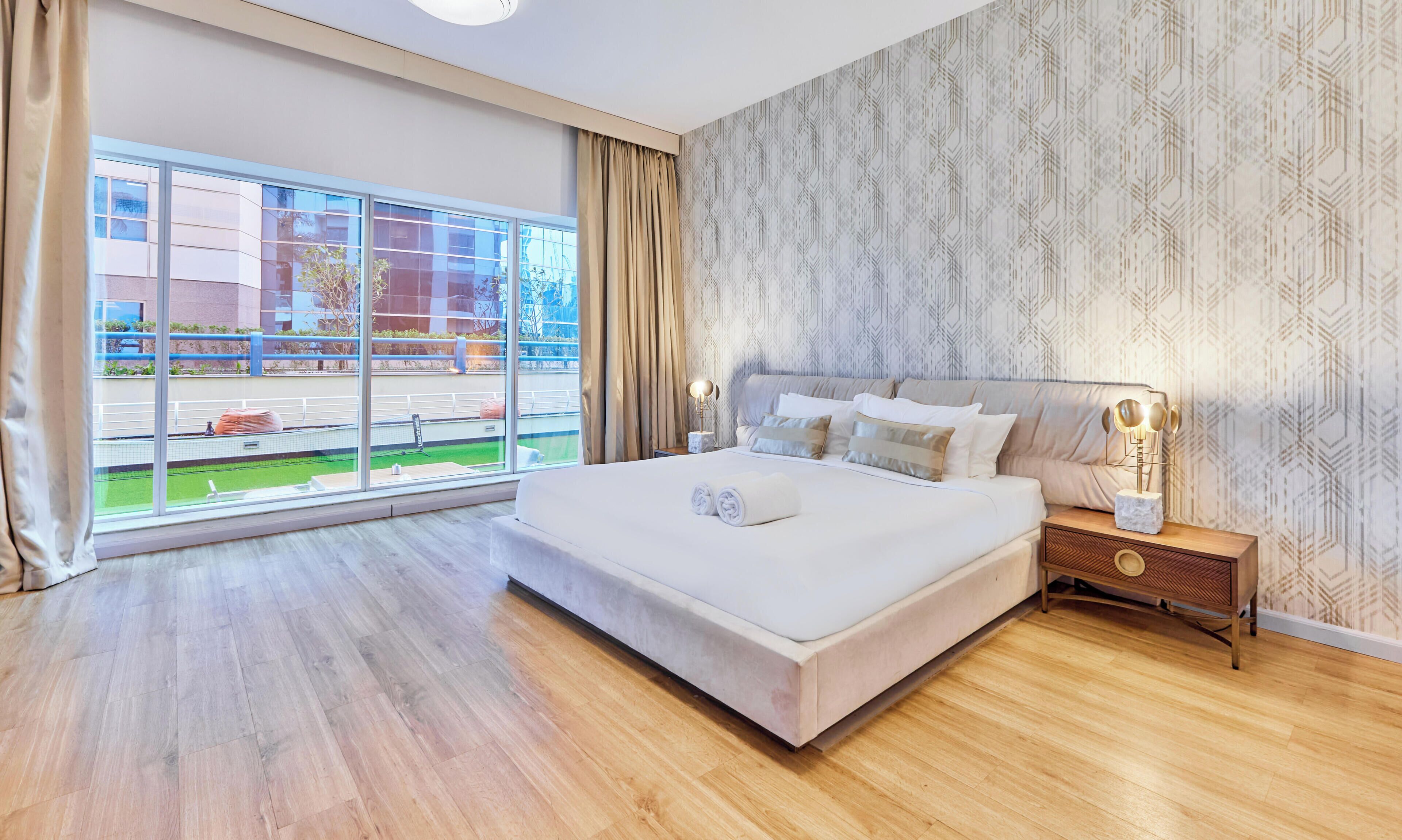 Camera d'albergo con un letto grande, un comodino con una lampada e vista su un campo da tennis, oltre a carta da parati decorata.