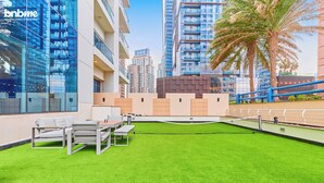 A modern outdoor area with a tennis court, seating, and high-rise buildings in the background. - bnbme - Emirates Crown - 2BR & Maid Room (Dubai)