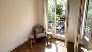 Una habitación con piso de madera, un sillón gris, un taburete pequeño y un ventanal corredizo que da a un balcón exterior con vistas a edificios y árboles.