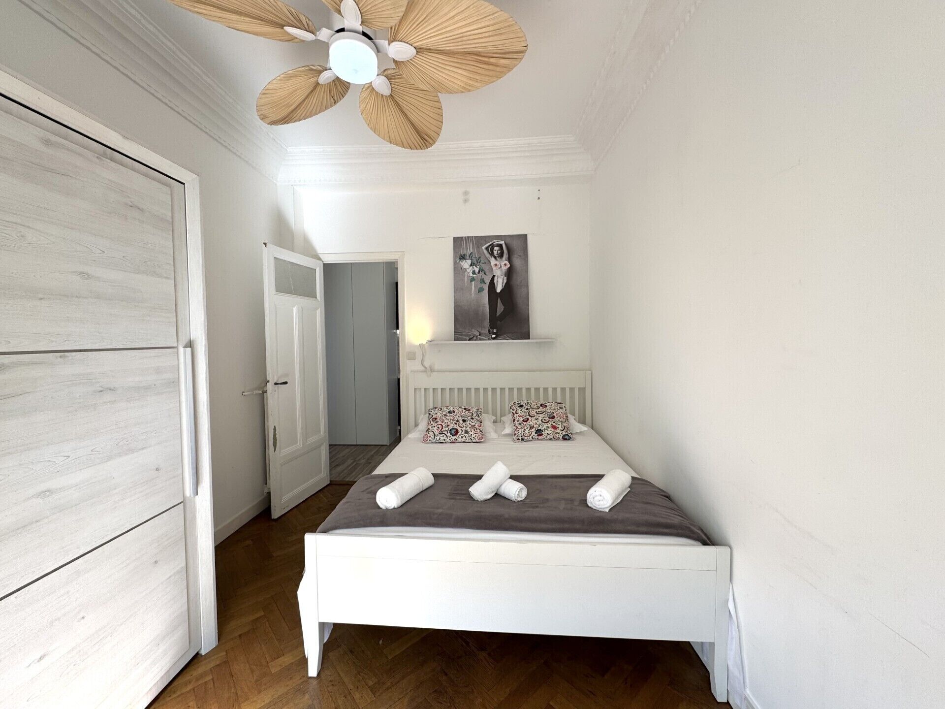 A bedroom with a white bed, a ceiling fan, a door, and a framed picture on the wall.