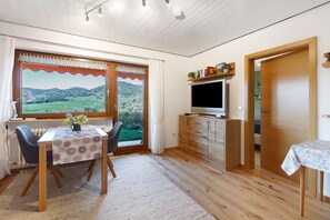 A room with a dining table, chairs, a TV, and a view of a green landscape through a window. - Apartment Rosi with Unique Panoramic View, Large Sun Terrace and Wi-Fi (Bernau im Schwarzwald)
