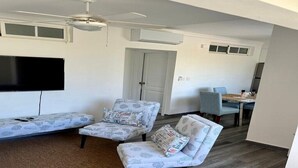 Ein Wohnzimmer mit einem Flachbildfernseher, zwei gemusterten Sesseln, einem Deckenventilator und einem Essbereich mit Tisch und Stühlen.