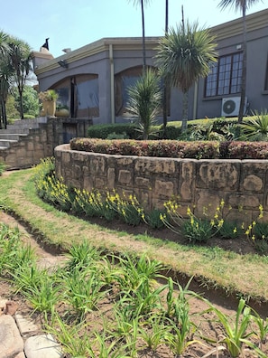 Un jardín ajardinado con muro de piedra, flores amarillas y un edificio de techo plano y ventanas.