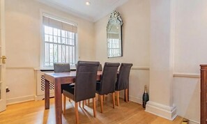A dining area with a wooden table and four chairs, a large mirror on the wall, and a window with bars. - Premium Central London Stay Near Sloane Square (London)