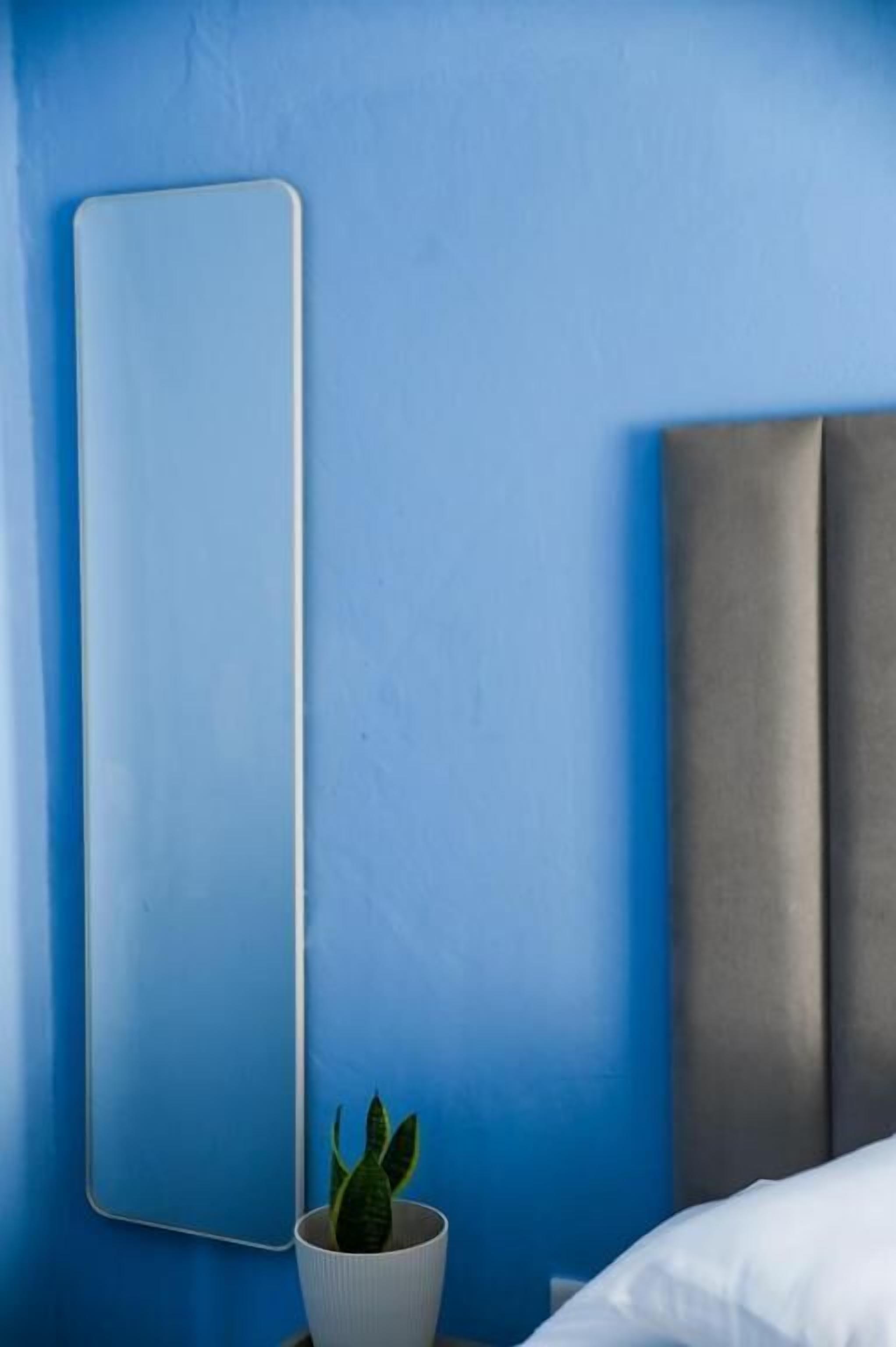 Un dormitorio con pared azul, una planta en la mesita de noche, un almohadón blanco y un cabecero.
