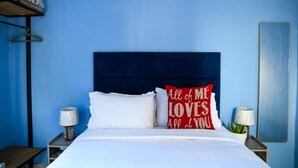 Habitación de hotel con pared azul, cabecero oscuro, ropa de cama blanca y almohada roja con texto.