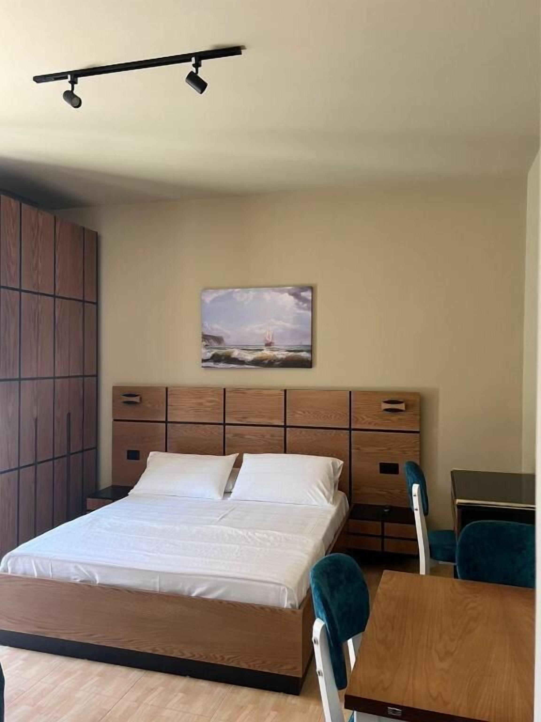 A bedroom with a bed, a wooden headboard, a painting, a desk, and a chair.